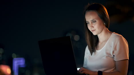 A-young-woman-on-a-summer-night-sitting-on-the-grass-in-the-city-looks-at-the-laptop-screen-and-writes-a-message-on-the-keyboard.-Programmer-freelancer.-Remote-night-work